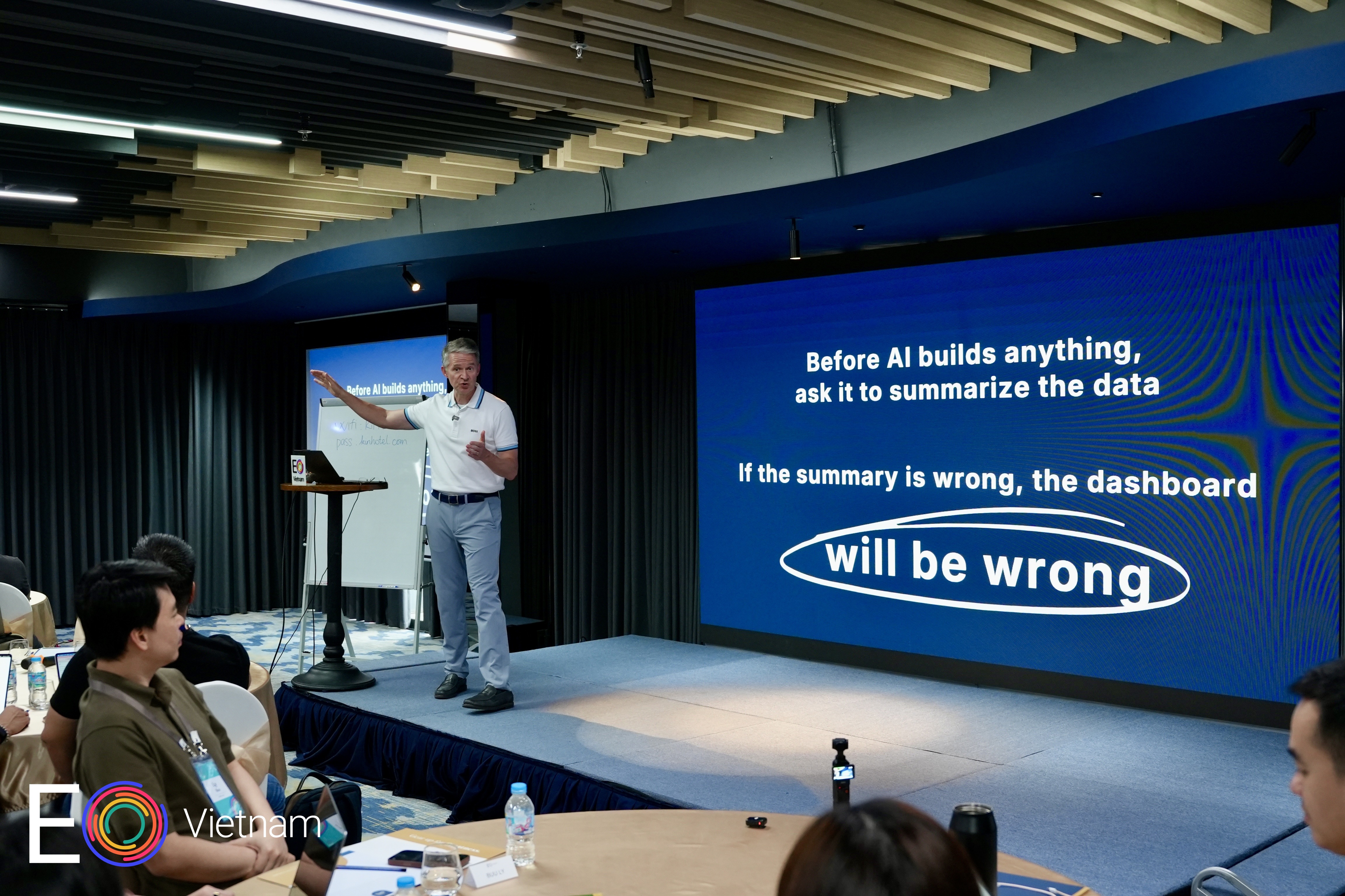 Brooks C. Holtom presenting at EO Vietnam, standing on stage beside a slide reading "Before AI builds anything, ask it to summarize the data"